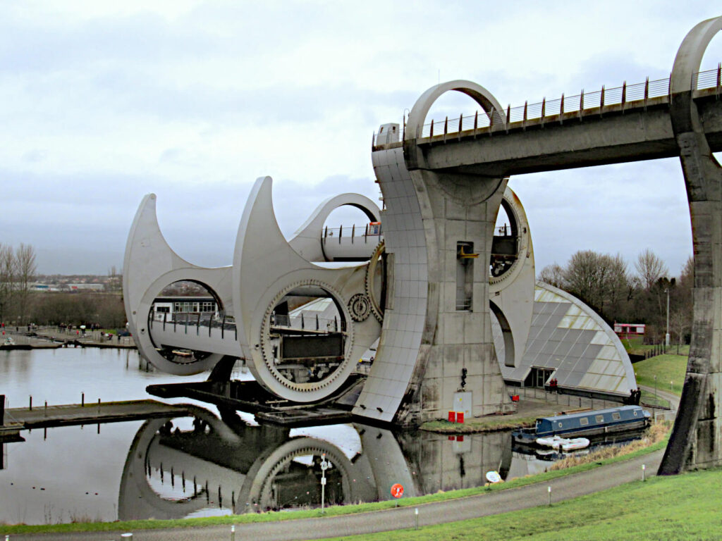 The Falkirk Wheel, a rotating boat lift, is shown mid-rotation. The lake next to it is very still and there are very few people around.