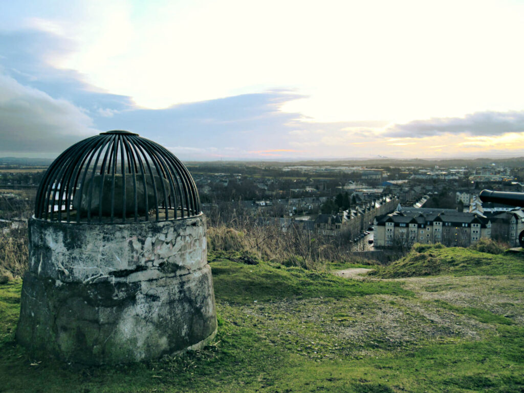 The beheading stone, the stone on top of the large stone to the left of the picutre, overlooks Stirling and the countryside beyond. Rows of houses continue into the horizon, and the sun is just rising into a cloudy sky.