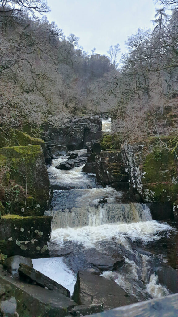 Water rushing down the Bracklinn Falls, white water is created from the amount of rocks. Bare trees line each side of the water.