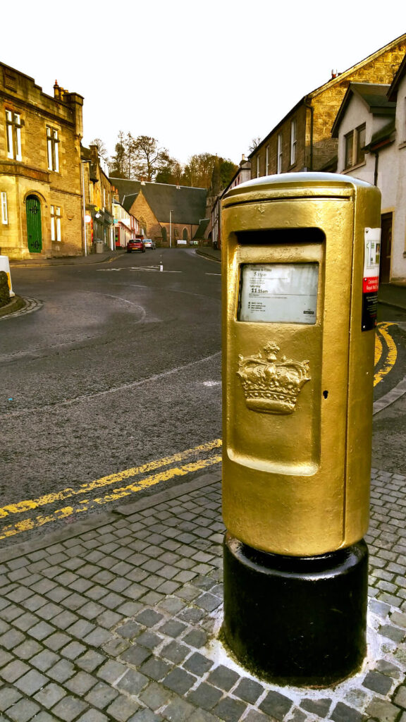 Golden post box. Painted in 2012 by Royal Mail to celebrate Andy Murray's Olympic Gold Medal in the Tennis Men's Singles. It is shown with houses and shops in the background on a quiet street.