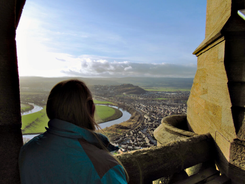 Zoe looking out over Stirling and Stirling Castle from the lookout at the top of the Wallace Monument. The sun has just risen, the light is golden and the sky is mainly blue with some clouds in the distance. 