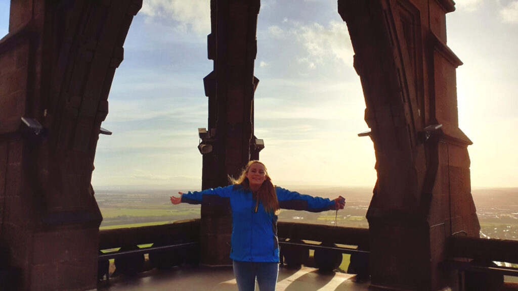 Zoe stood at the lookout of the Wallace Monument. The sun is low in the sky and the light is golden and radiating. Zoe is wearing a blue coat with both arms outstretched horizontally. 