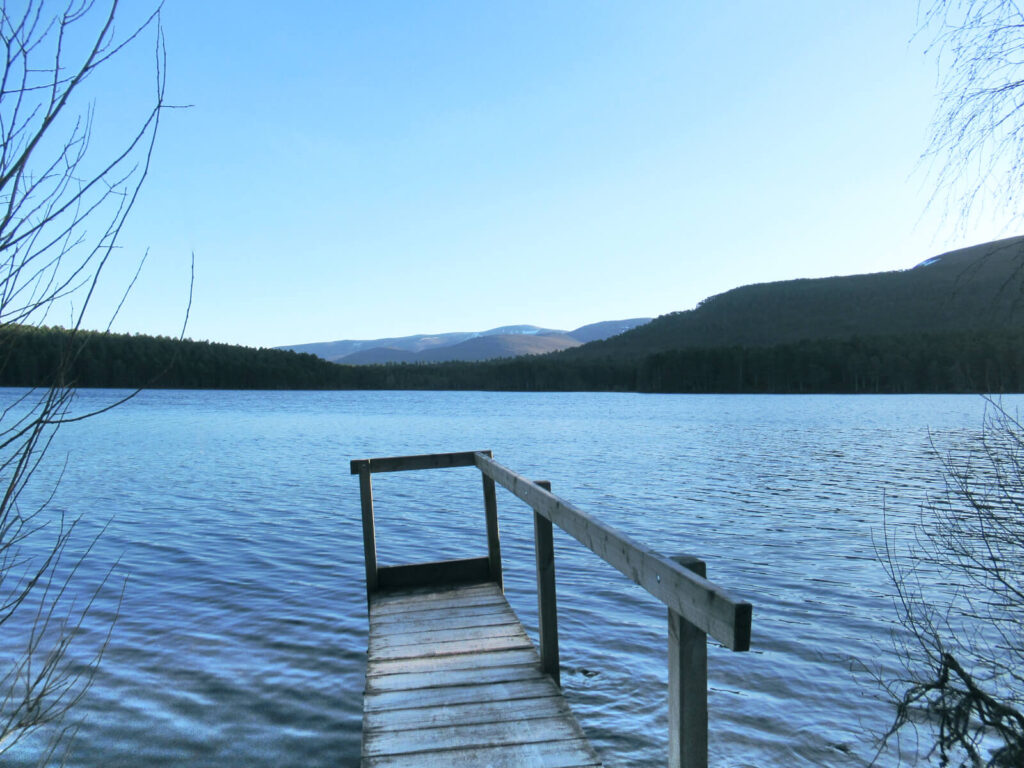 A small jetty extends into the loch, which is a rich blue colour and slightly wavy. There are wooded mountains in the background with a small layer of snow at the top. The sky is crisp blue