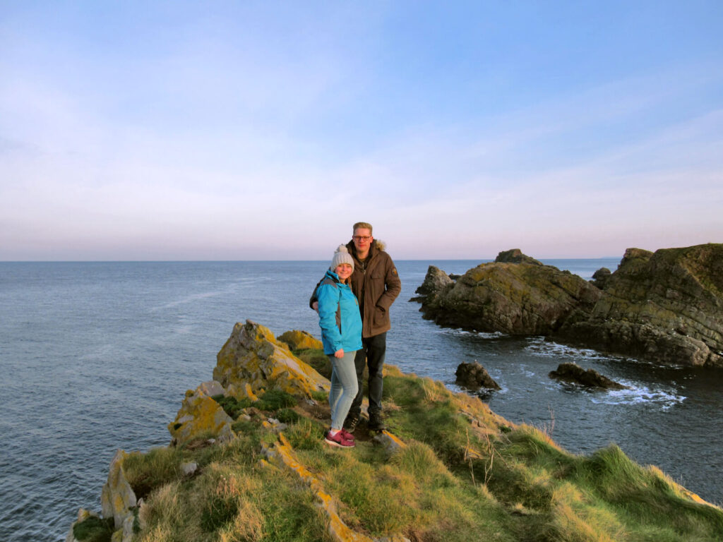 Zoe and boy-Zoe stood on a cliff top overlooking the Moray Firth. It's just before sunset and there are pink tinges to the sky on the horizon. Waves crash against the rocks that stick out from the sea just away from the coast line.