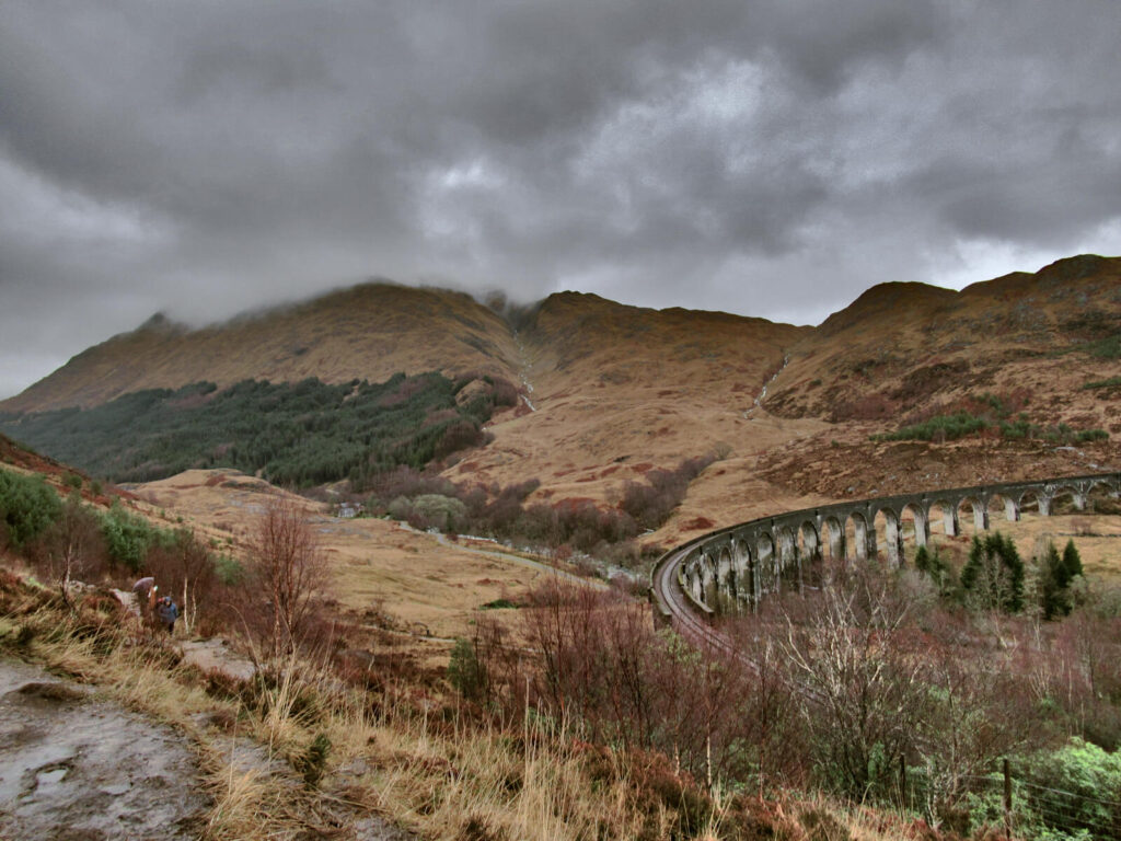 Dark grey clouds hang low on the hilltops behind the Glenfinnan Viaduct. The curve of the viaduct is on the right hand side of the picture and looks small in comparison to the surrounding hills. 