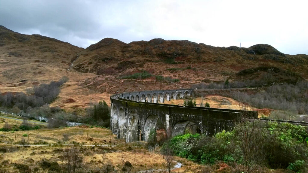 The curve of the Glenfinnan Viaduct is visible against the brown hills of Scotland and the grey skies