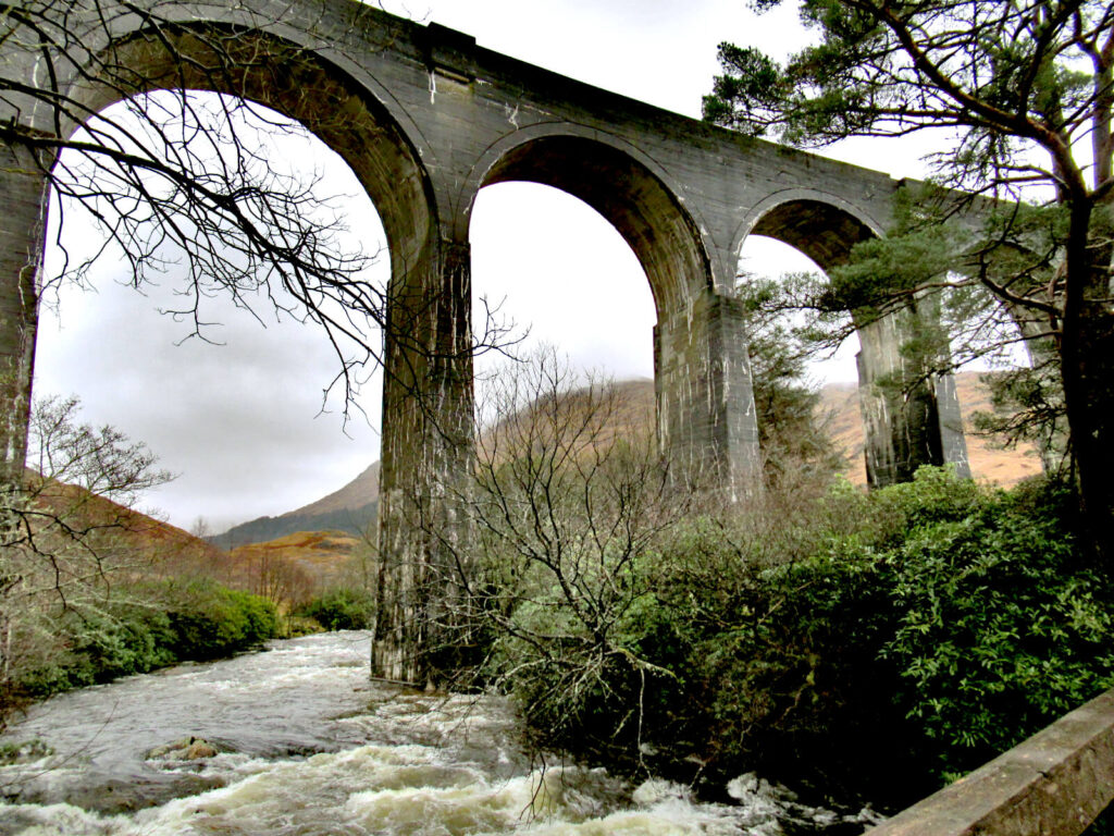 A river flows rapidly underneath the grey arches of the viaduct. Grey clouds can still be seen in the distant sky.