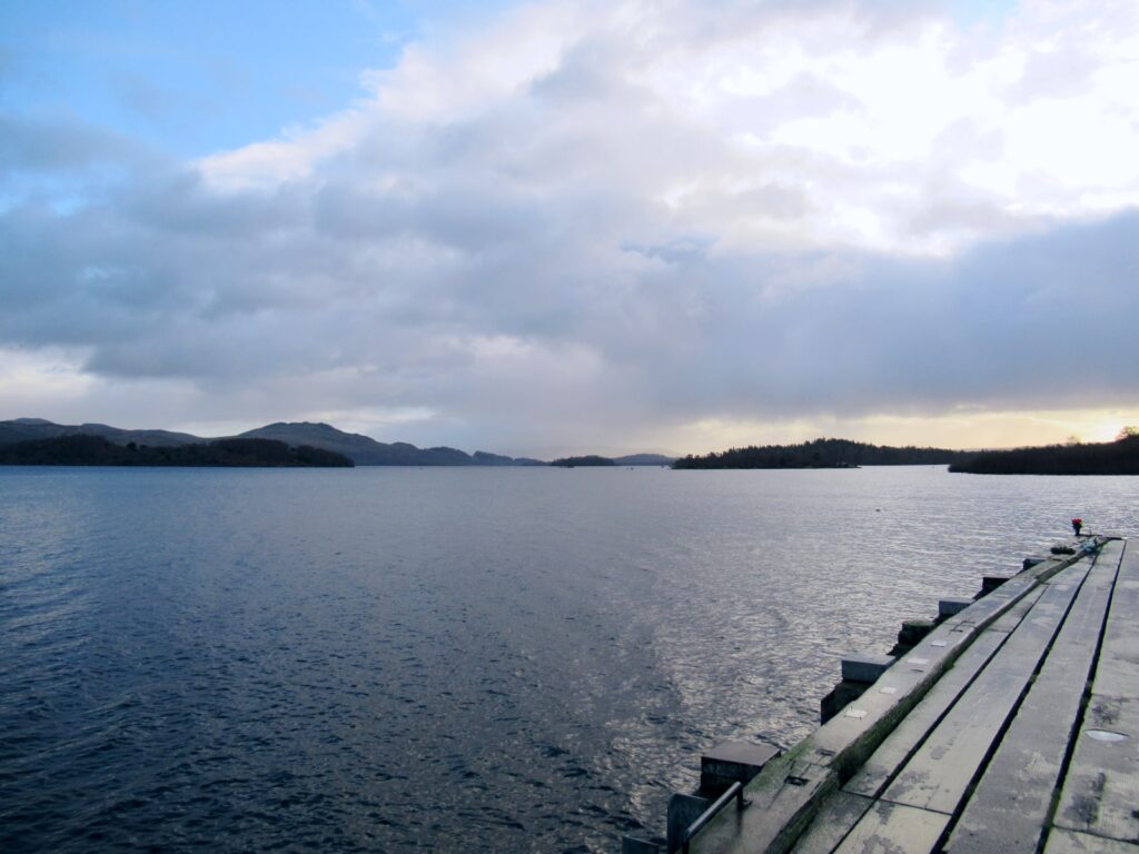 A cloudy day at Loch Lomond. The edge of a wooden jetty can be seen in the picture foreground and the water continues into the horizon. Small islands can be seen.