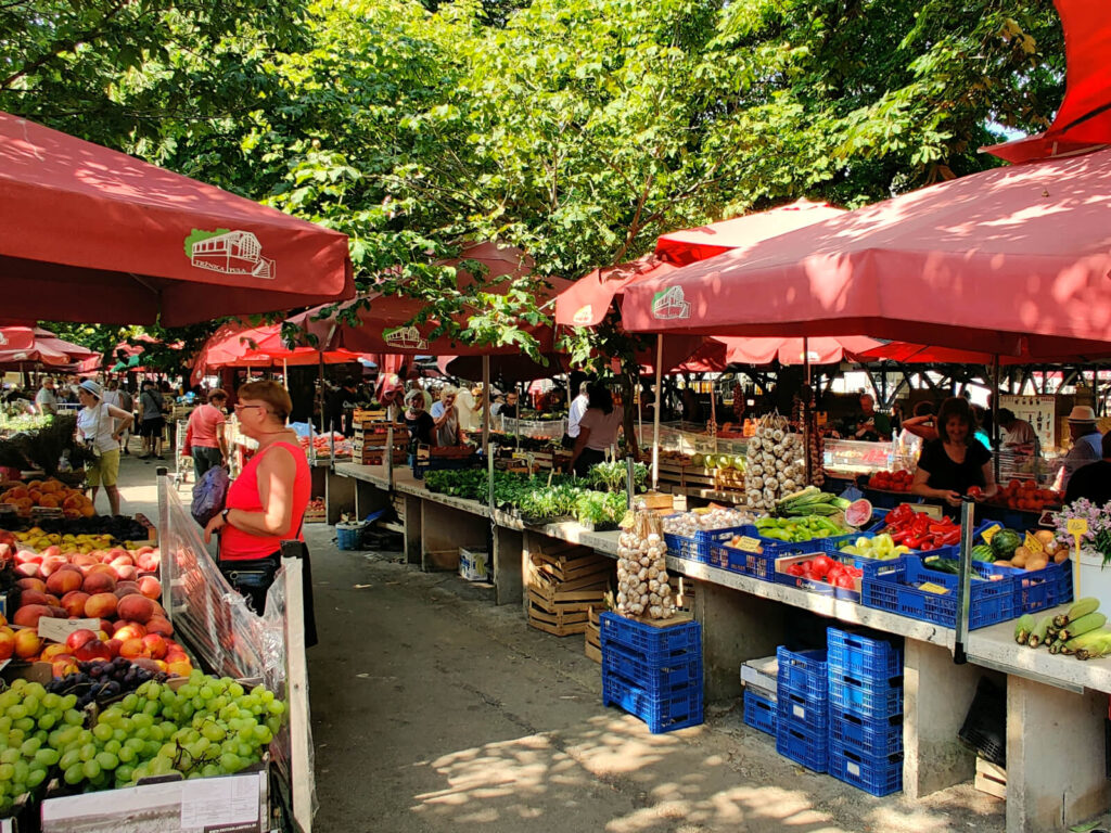 The photo shows a market scene, the stalls run in parallel lines and are sheltered by red covers. Fruit and vegetables of all colours are on display. Istria and Pula are worth visiting for the amazing food, with both Italian and Balkan influence!