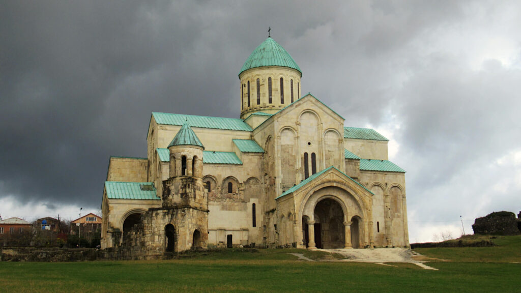 Bagrati Cathedral, with it's iconic teal green roof stands overlooking Kutaisi from the hill top. Dark rain clouds loom in the background but do not distract from the brightness of the building, which has a sandstone colour.