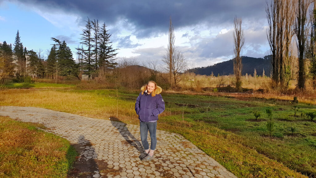 Zoe stood in Kutaisi Botanical Garden during golden hour, the light is low and warm. Winter trees are all around
