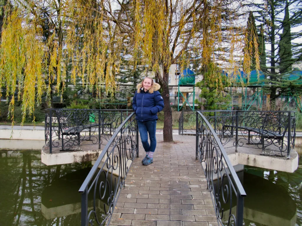 Zoe stood on a a bridge over a pond in Kutaisi Botanical Garden. A weeping willow tree hangs low with its winter yellow leaves.