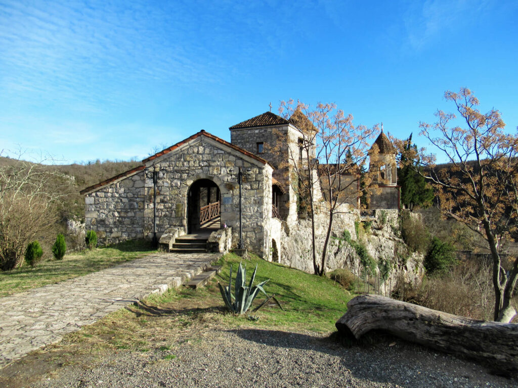 The Motsameta monastery: an old stone building set into the side of a hill surrounded by lush green land. The winter sky is clear and crisp blue.
