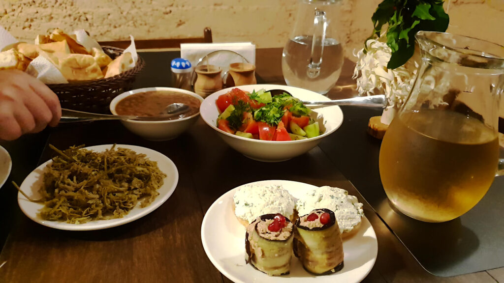 A selection of Georgian cuisine on a table including a jug of amber wine, a bowl of salad, cream-cheese-like substance on crackers and a bowl of bread in the background