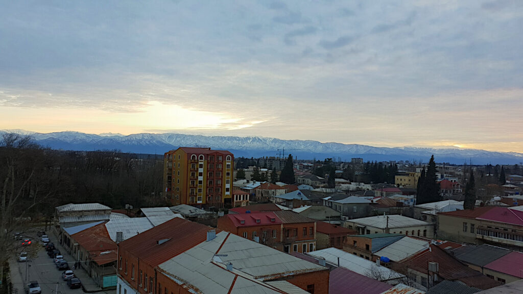 Looking south from Kutaisi towards the snowy mountains at sunset. The sun has just dipped below the mountains and the sky has a yellow tint. In the foreground the 1970s style buildings of Kutaisi can be seen