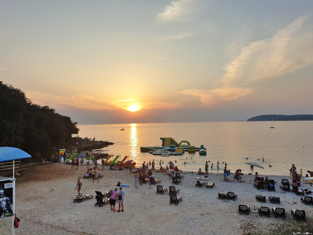 Ambrela Beach at sunset, people are dotted across the beach and shallow sea. The sun is golden and just above the horizon. One of the best beaches in Pula