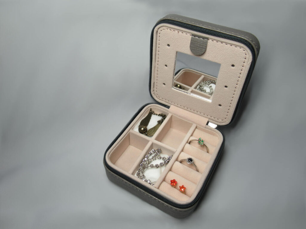 Small travel jewellery box shown containing a small mirror, two rings and a pair of ear rings in narrow cushioned partitions. Two bracelets are shown in a separate partition. Take inspiration for unique travel gifts!