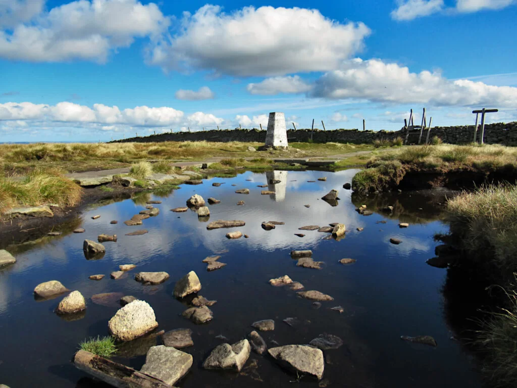 A beautiful day on the summit of Buckden Pike, blue sky and fluffy clouds. The reflection of the trig point in the puddle adds to the calm atmosphere