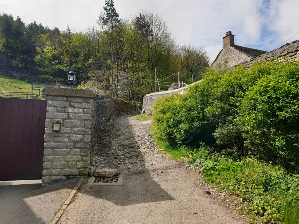 The start of the path from Castleton to Speedwell Cavern. The stoney path runs between a stone exterior wall and green bushes.