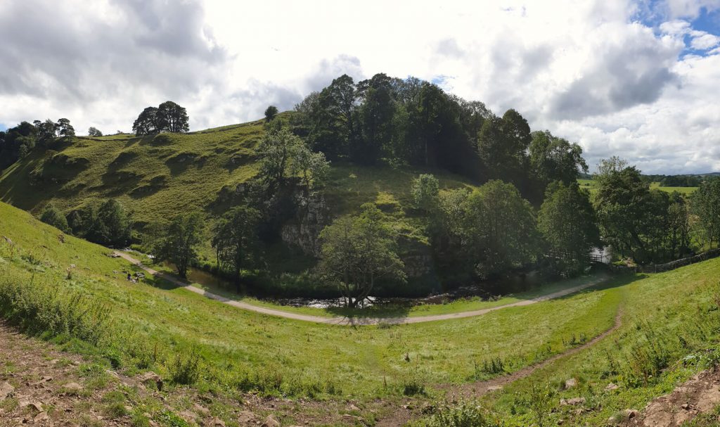 A panoramic shot of the start of Wolfscote Dale from inside a cave in the valley wall. The image shows the path and river at the bottom of the valley surrounded by lush green grass and lots of trees. This point is near the end of this Hartington Walk