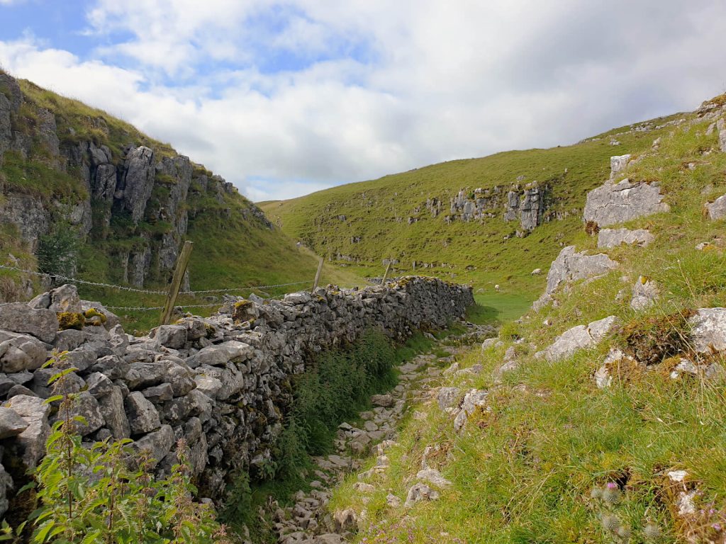The path runs parallel with a dry stone wall and between the cliff faces of rocky hills