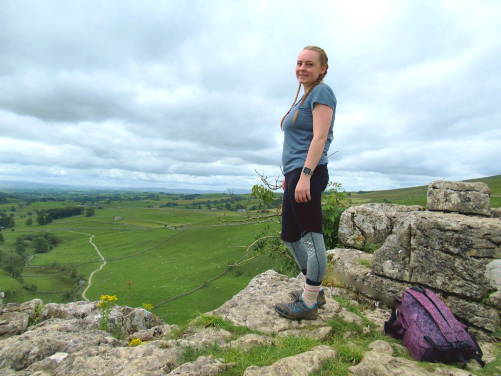 Zoe is stood on the edge of the top of Malham Cove - the first stop on this Malham Circular Walk. The view looks out over the Yorkshire Dales and there are a number of hills on the horizon.