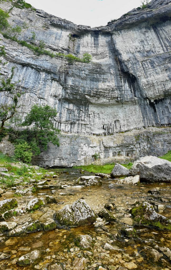 From the bottom of Malham Cove, water trickles out from under the rockface and along the strem. The limestone rocks rise high into the sky