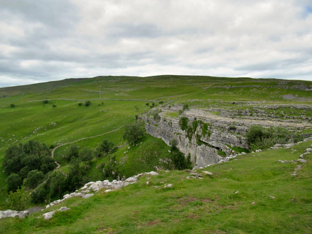 Taken from the far side of the top of Malham Cove, the rocks and cliff face are visible, the surrounding area is very green and the hill rises into the background