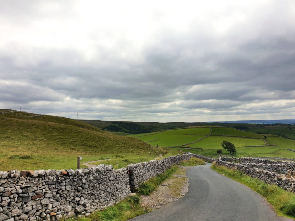 The photo is taken from the middle of a quiet, single-path country road. The road snakes off into the distance and empty fields and rolling hills can be seen in the distance. A dry stone wall runs parallel to the road on either side