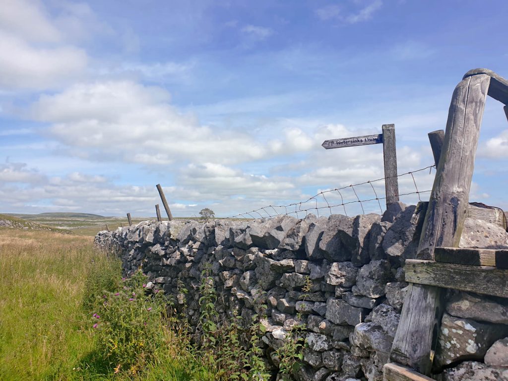 Dry stone wall with stile crossing from the road onto the footpath towards Watersinks Car Park. This marks roughly half way on the final leg of this Malham Circular Walk.