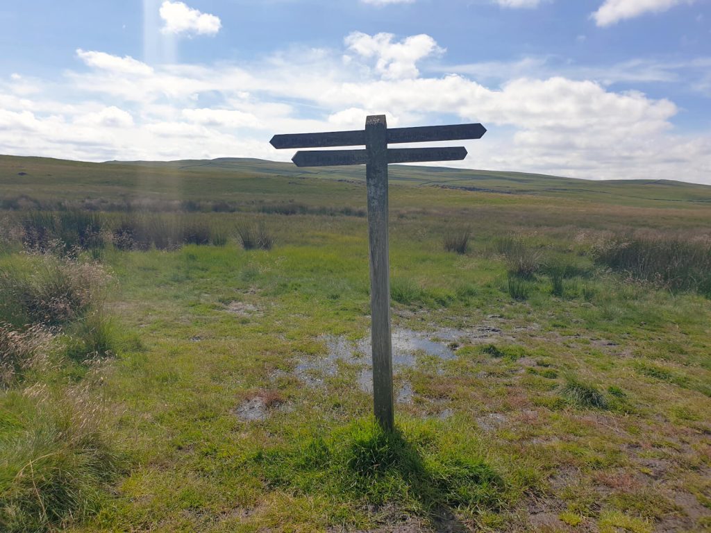 Almost at the end of the Malham Circular Walk! 4-way signpost in the middle of a green field with rolling hills in the background