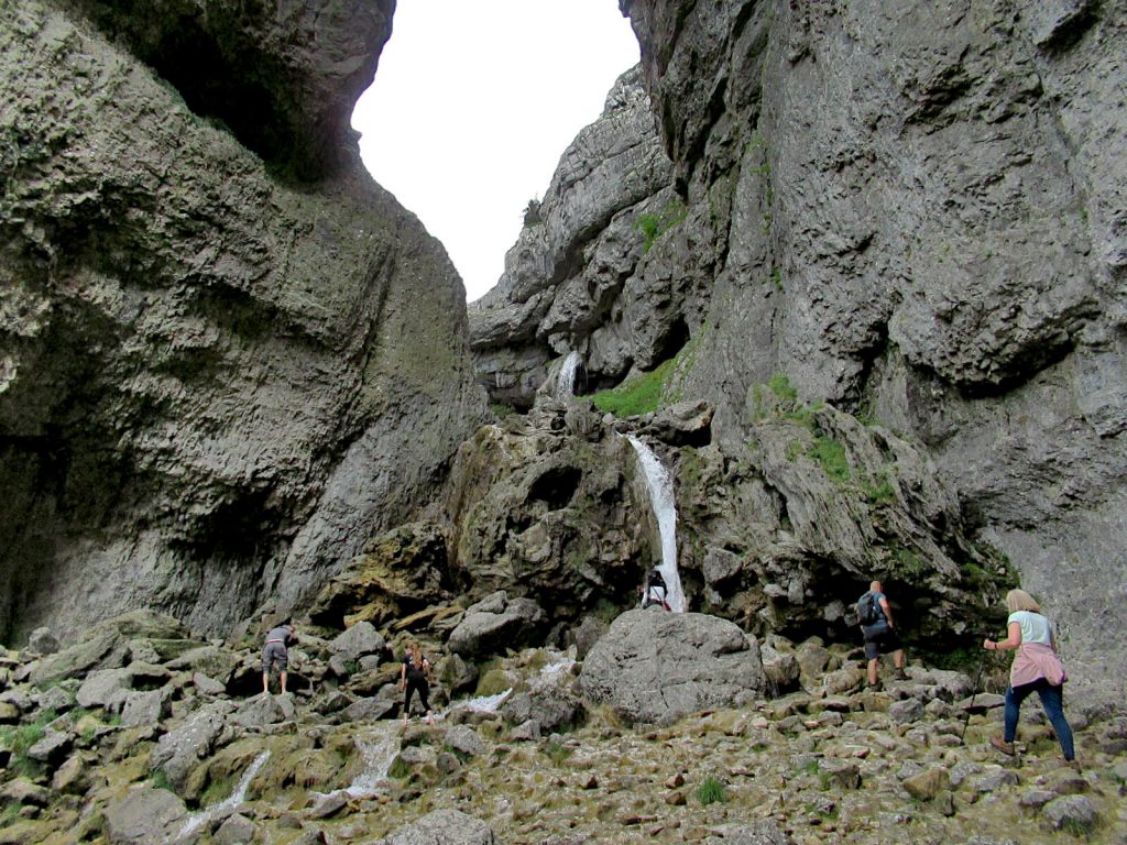 The Gordale Scar waterfall crashes over rugged limestone, brown rocks. A second waterfall is seen in the background. A few people are at the bottom admiring the natural sight. From here, one option on the Malham Circular Walk is to climb the left side of the waterfall to rejoin the path above. An alternative and less risky option is also available