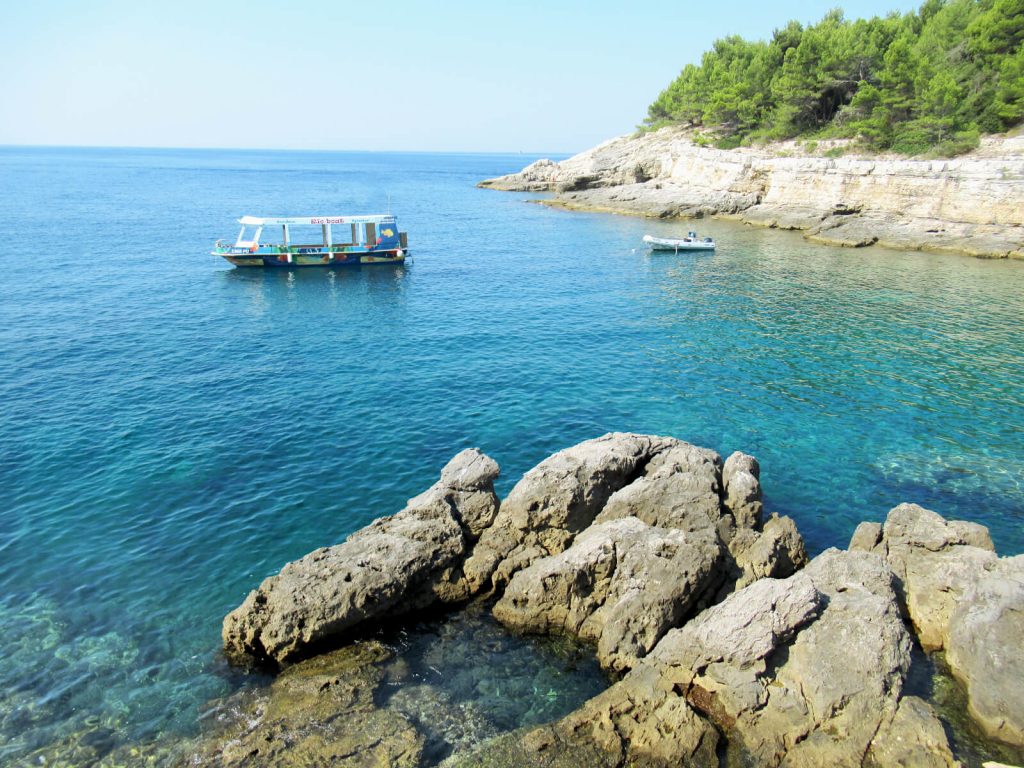 Photo taken from the top of a small cliff looking out over the bright blue sea. Rocks are visible in the foreground as they emerge above the waterline. Two boats are anchored in the water and a forested coastline falls into the sea in the background