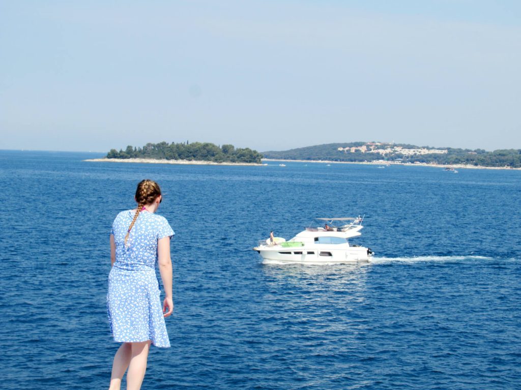 Zoe stood in a blue and white dress facing away from the camera and looking out to sea. A small, white yacht is passing by surrounded by the dark blue sea. In the background a small island with lots of trees is visible. The sky is blue and there are no clouds in sight! One of the best things to do in Pula is to take a trip to Rovinj!
