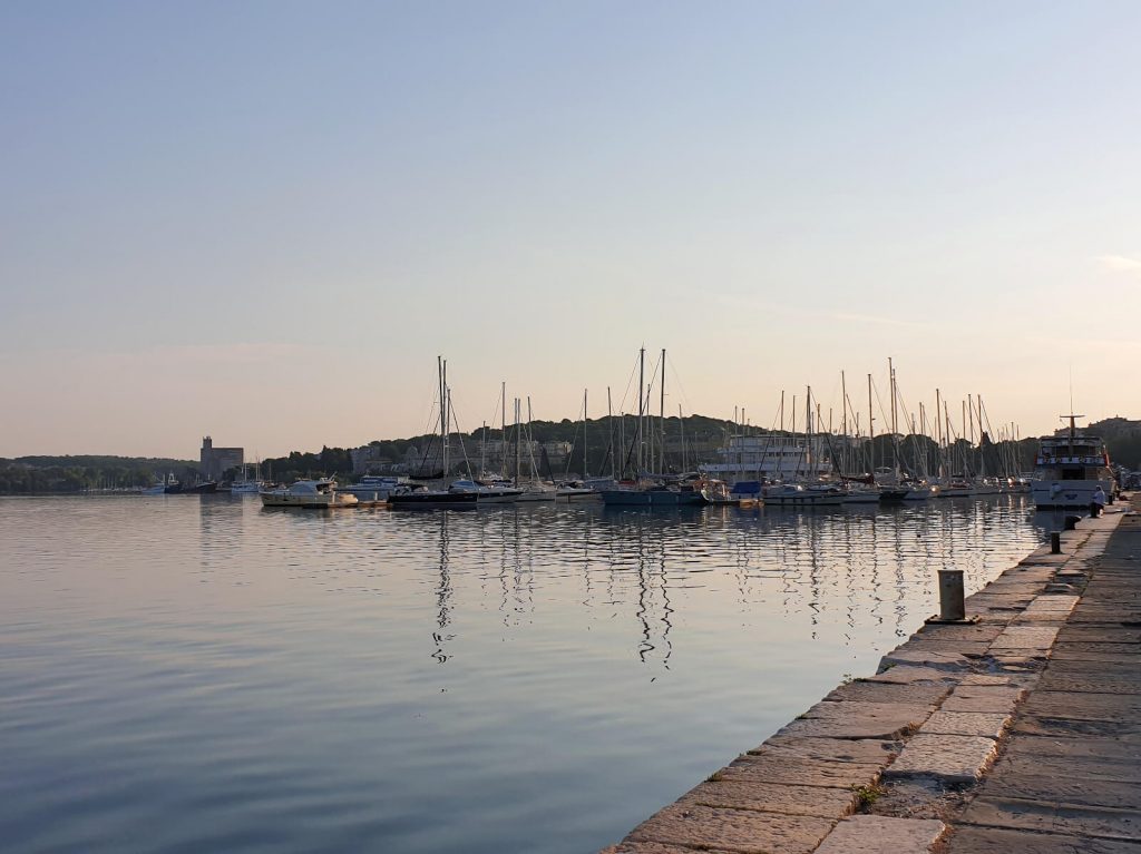 Sail boats without masts line Pula Marina. It is just after sunrise and the light is faint and soft. The water is calm and the sky is clear.