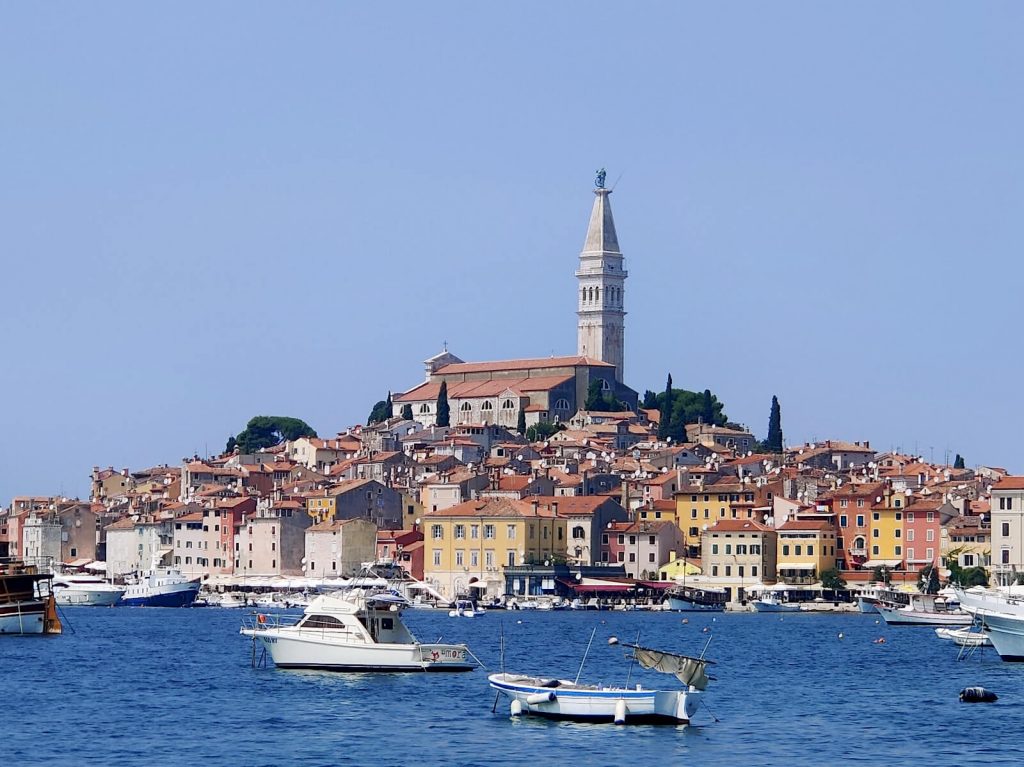 The church tower stands above the other red-roofed buildings that make up Rovinj's old town. In the foreground many boats are docked in the marina. The colourful buildings contrast with the bright blue sea. Taking a day trip here is one of the best things to do whilst in Pula!