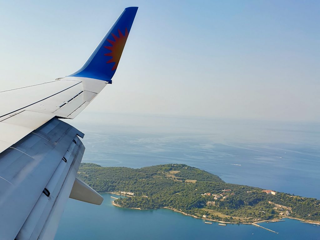 Taken from the window seat of a Jet2 plane on the approach to Pula from the west. You can see a white wing, with the flaps deployed with a blue tip containing half a sun (cut vertically down the middle). On the ground you can see a peninsula of Pula, with lots of forest and surrounded by the blue sea that goes into the horizon