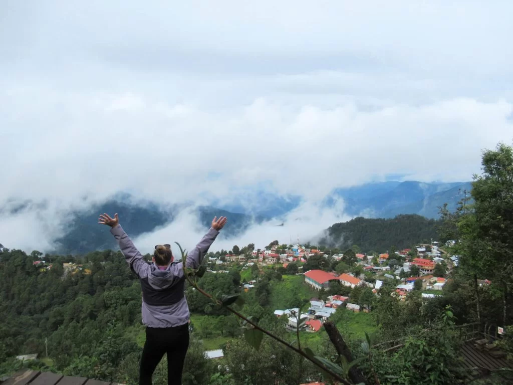 There are many things to do in San Jose del Pacifico including climbing to this mirador for impressive views. Zoe is stood looking away from the camera with both arms above her head. The clouds cover the mountains in the distance