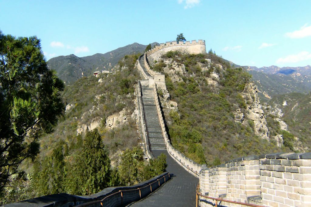 An uphill stretch of the Great Wall of China, visitable from Beijing on a daytrip. Either side is lush green trees and grass