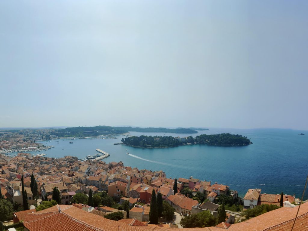View of the Adriatic Sea and southern Rovinj from the Bell Tower. The orange roofs of the town can be seen in the foreground and a small island sits in the middle of the sea in the centre of the photo.