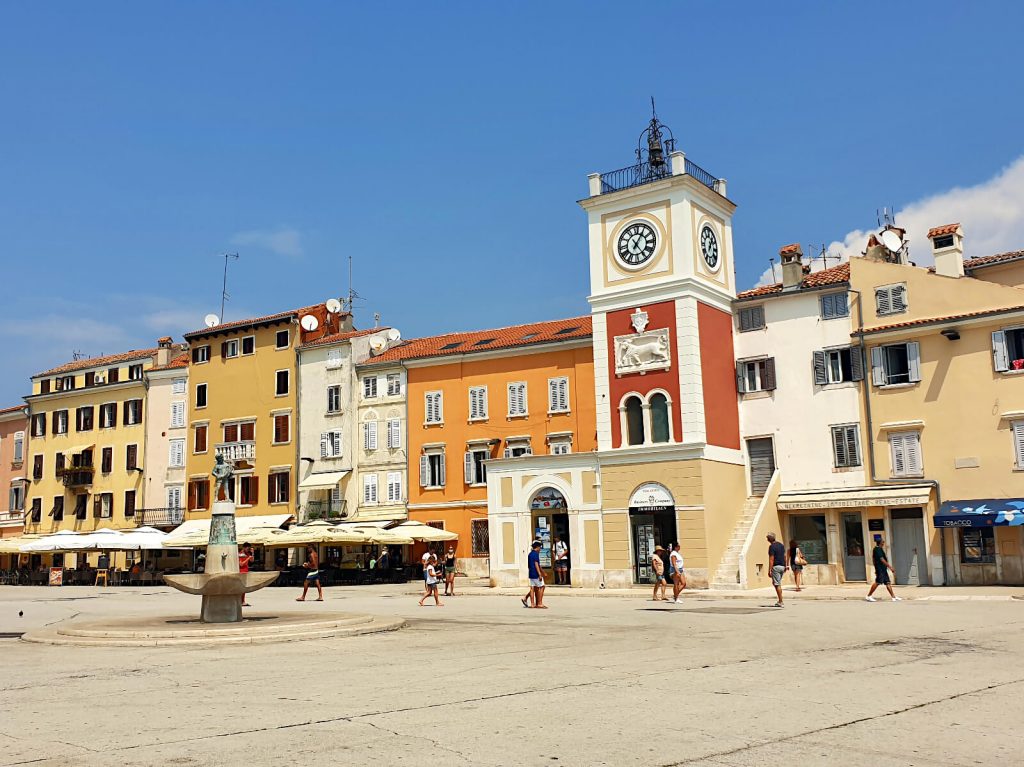 The square in the centre of Rovinj. There is a large open space iwth a statue in the middle, surrounded by buildings that are orange, yellow and beige.