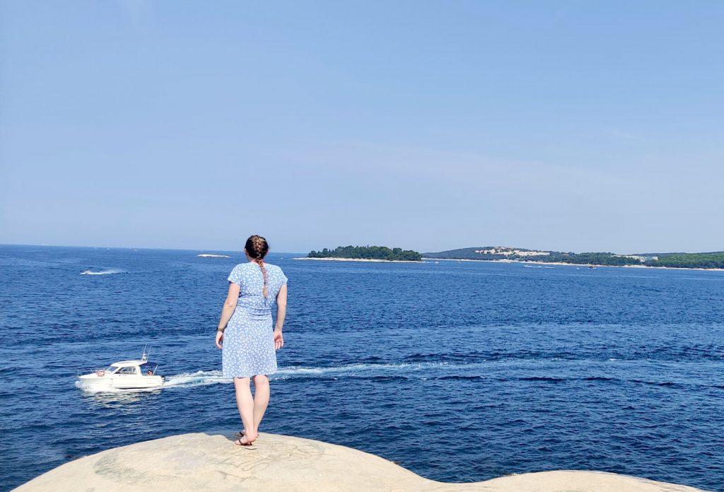 Zoe stood on top of an abandoned bunker looking out over the dark blue sea. Zoe is wearing a light blue and white dress with flip flops.