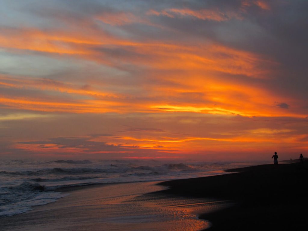 A vivid sunset over the Pacific Ocean. The sky is various shades or blue and orange. A single silhouette stands on the beach